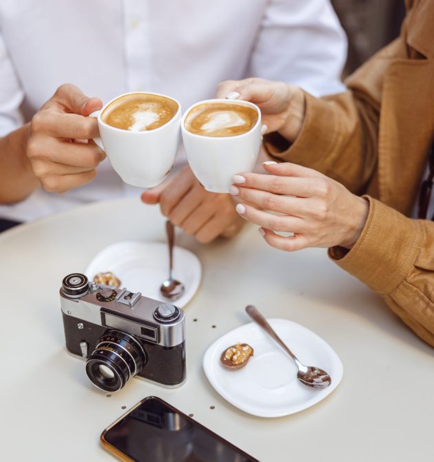 Woman and man holding cups of latte with heart pattern over table with camera and phone on it